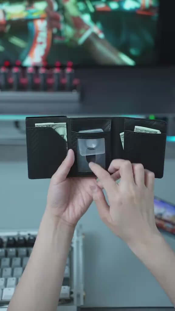 Hand holding a sleek black wallet 👜 with cash 💵 visible, on a desk with a computer set-up 💻.